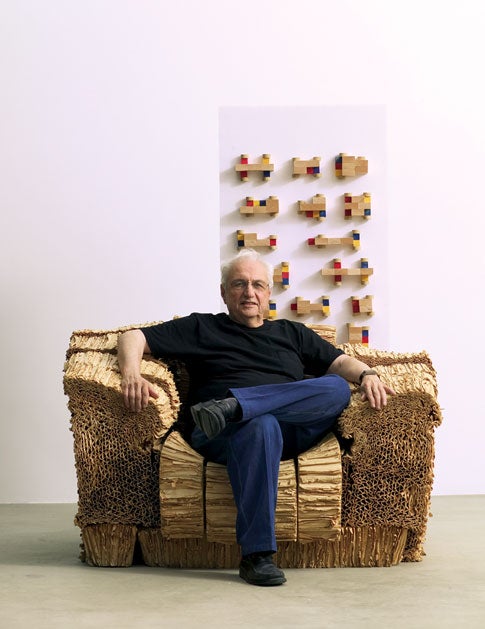 Architect Frank Gehry in a cardboard chair of his own design. Gehry is collaborating with a group of MIT students to make a new type of concept car. The colorful wood-block models behind him were built by one of the students.