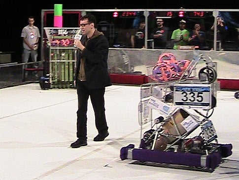 Fred Armisen in the center of an arena in front of a crowd at the 2009 FIRST Robotics Competition in New York City.