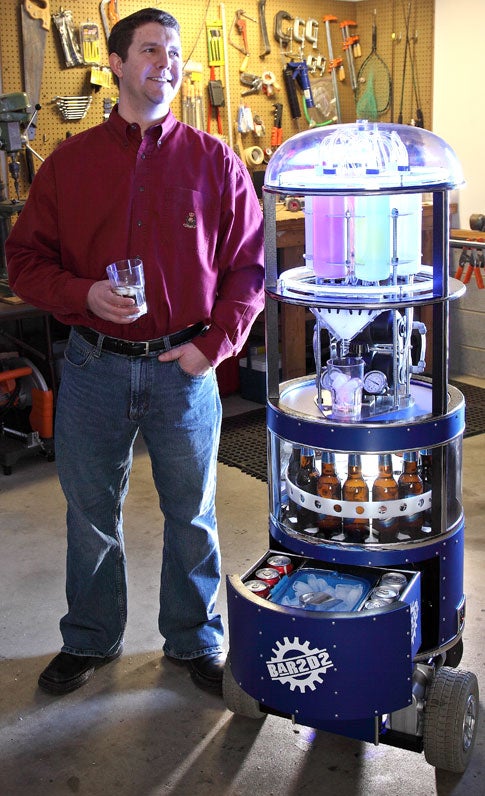 Jamie Price, wearing a red dress shirt and jeans, holding a beer next to his homemade bartending robot named Bar2D2.