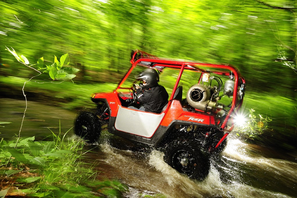 A man wearing a black firesuit and black helmet driving a jet turbine-powered ATV through a stream in the woods.