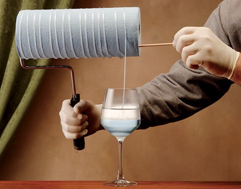 A person holding a paint roller with a roll of paper towels on it and using it to spin nylon thread out some some chemicals in a wine glass.
