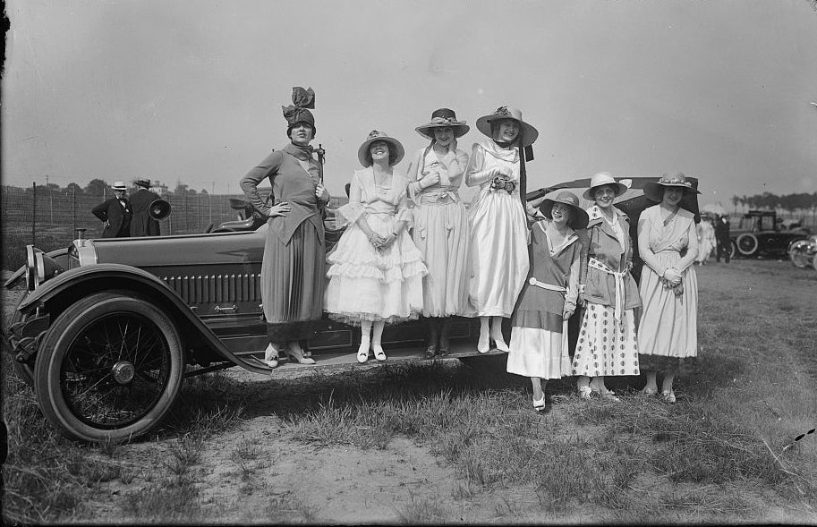 A group of well-dressed white women stand on a car, looking very 1910.