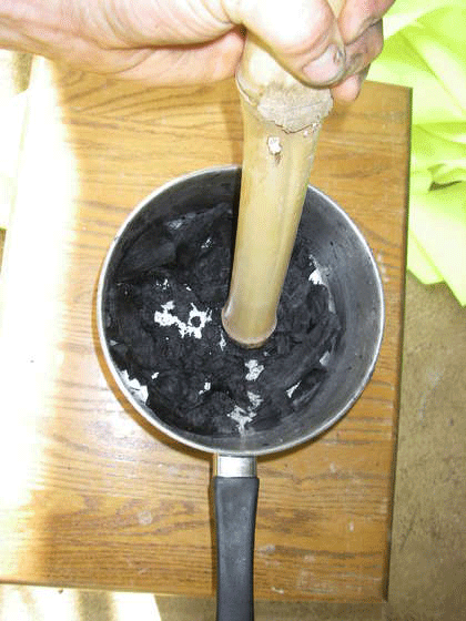 After quenching the coals I set them on some cardboard to drain and dry out a little. Then I dumped the water out of the pot and used it for a mortar to crush the charcoal in. I used a section of bamboo as a pestle. Quenching had drawn a lot of water into the charcoal. It turned to wet paste as I crushed it. I mashed it til the largest grains were well under .25" in diameter. The volume decreased a lot as I crushed it. This husk charcoal wasn't very dense. I needed to grind a few batches to get enough to fill my canister.