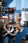 A man, Mike Begley, holding a DIY drag racer that's half circular saw and half rocking horse.