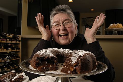 A white woman with short black-and-gray hair looking excited behind a chocolate bundt cake with a large slice taken out of it.