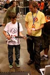 A man in a yellow t-shirt watching a young boy play with a modified Guitar Hero controller at Maker Faire 2008 in San Francisco.