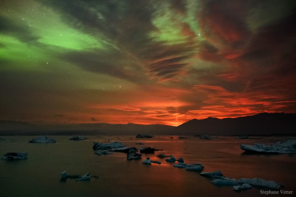 A rare volcanic light pillar, seen in the background of this image, shines in Iceland. The pillar is caused by glowing lava reflecting its light on ice in the atmosphere. Above, a green aurora shimmers in the sky.