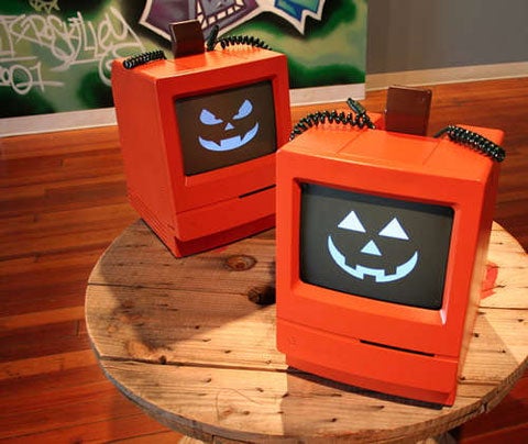 Two small orange computers with digital jack o' lantern faces on them, sitting on a wooden stool.