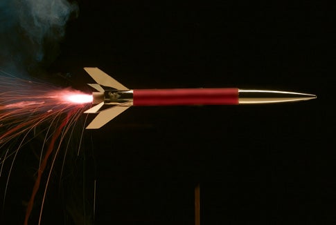 A model rocket with a red body and gold tip and fins, with flames coming out the back, against a black background.