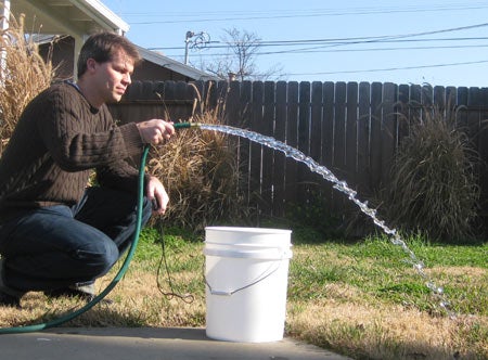 A person spraying water out of a garden hose over a white 5-gallon bucket in a fenced-in yard.