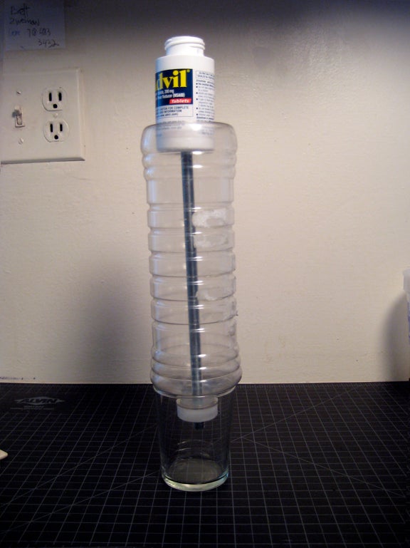 A plastic bottle upside down on top of a small glass with a metal rod inside the plastic bottle and an Advil bottle on top of it all.