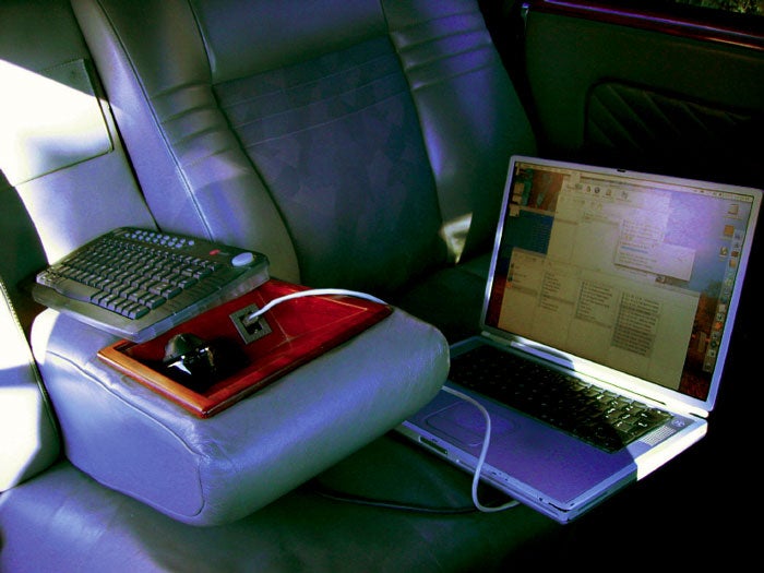 A Mac laptop on the seat of a car, connected to a keyboard and the car itself.