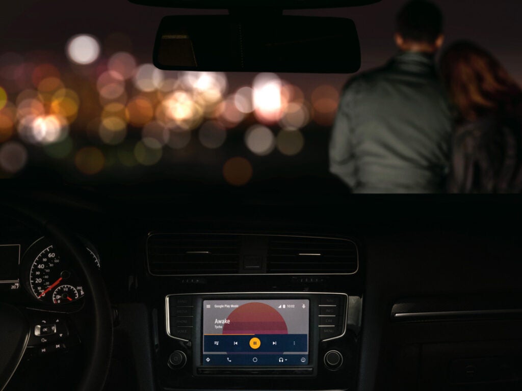 A night scene with a car dashboard in the foreground and a man and a woman sitting on the car hood in the background, city lights in the distance