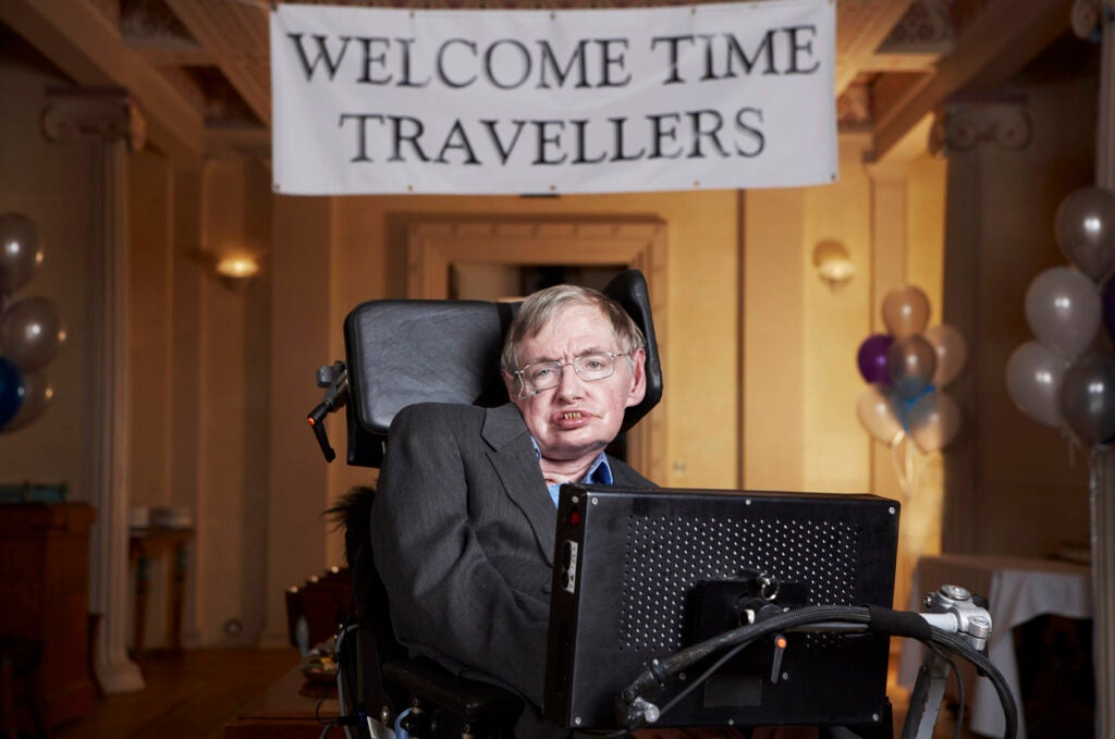stephen hawking sits in front of a sign that says "Welcome Time Travelers"