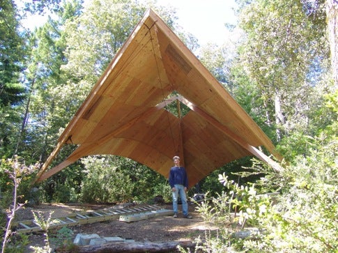 A man wearing a blue shirt and jeans stands under a wooden conical shelter.