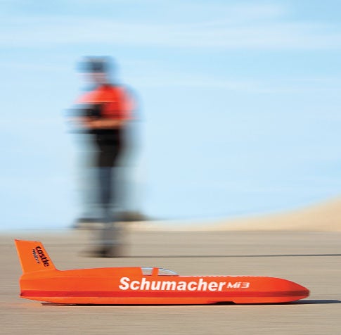 An orange rocket-shaped remote-control car with Schumacher written on the side in white text. There is a blurry person in the background as the car passes by on flat desert terrain.