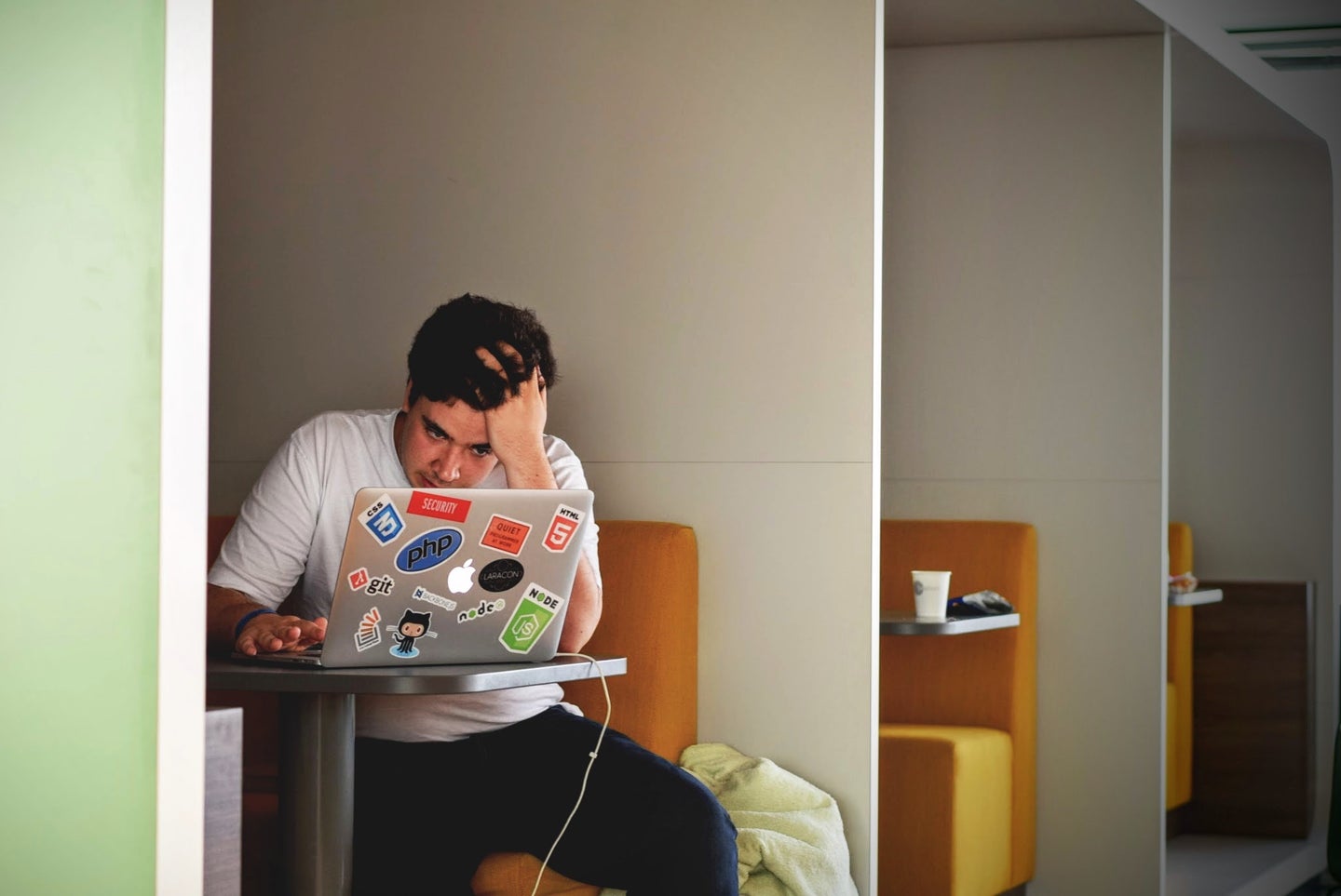 A man sitting in a study cubicle, holding his head in frustration as he looks at his laptop, which has a lot of stickers on it.