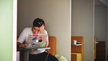 A man sitting in a study cubicle, holding his head in frustration as he looks at his laptop, which has a lot of stickers on it.