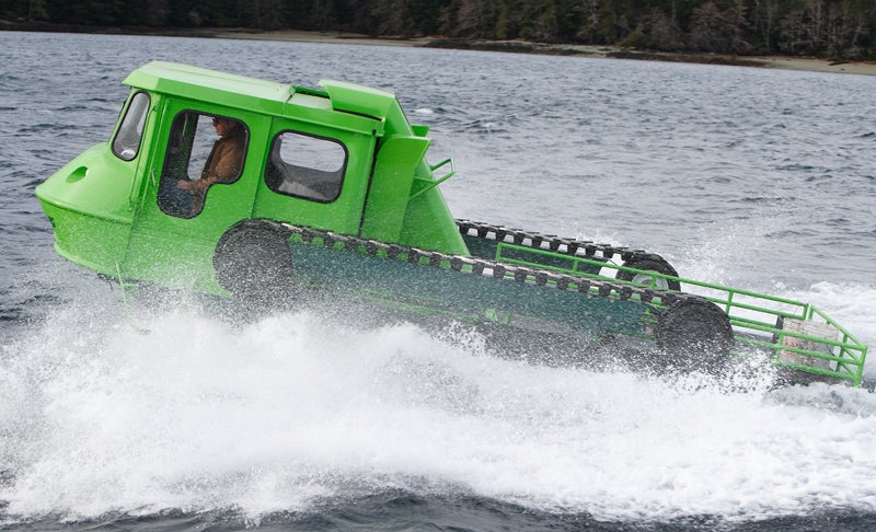 A green amphibious vehicle splashing through waves on a body of water.