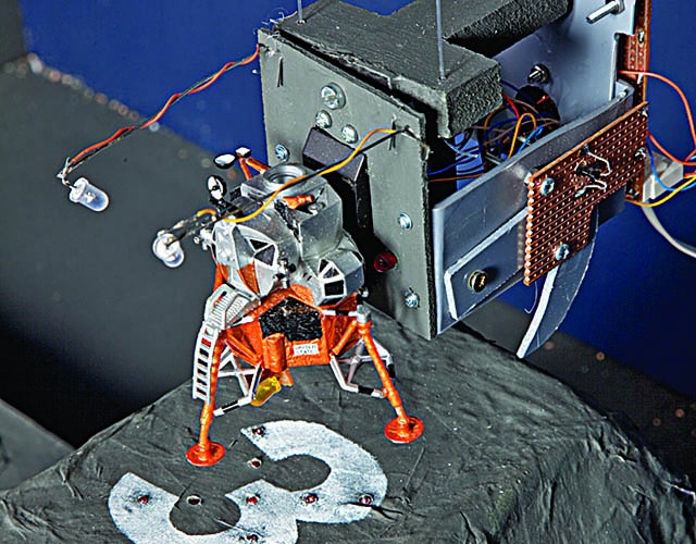 A model lunar lander connected to an arm that holds and controls it, above a landing zone with the number "three" on it.