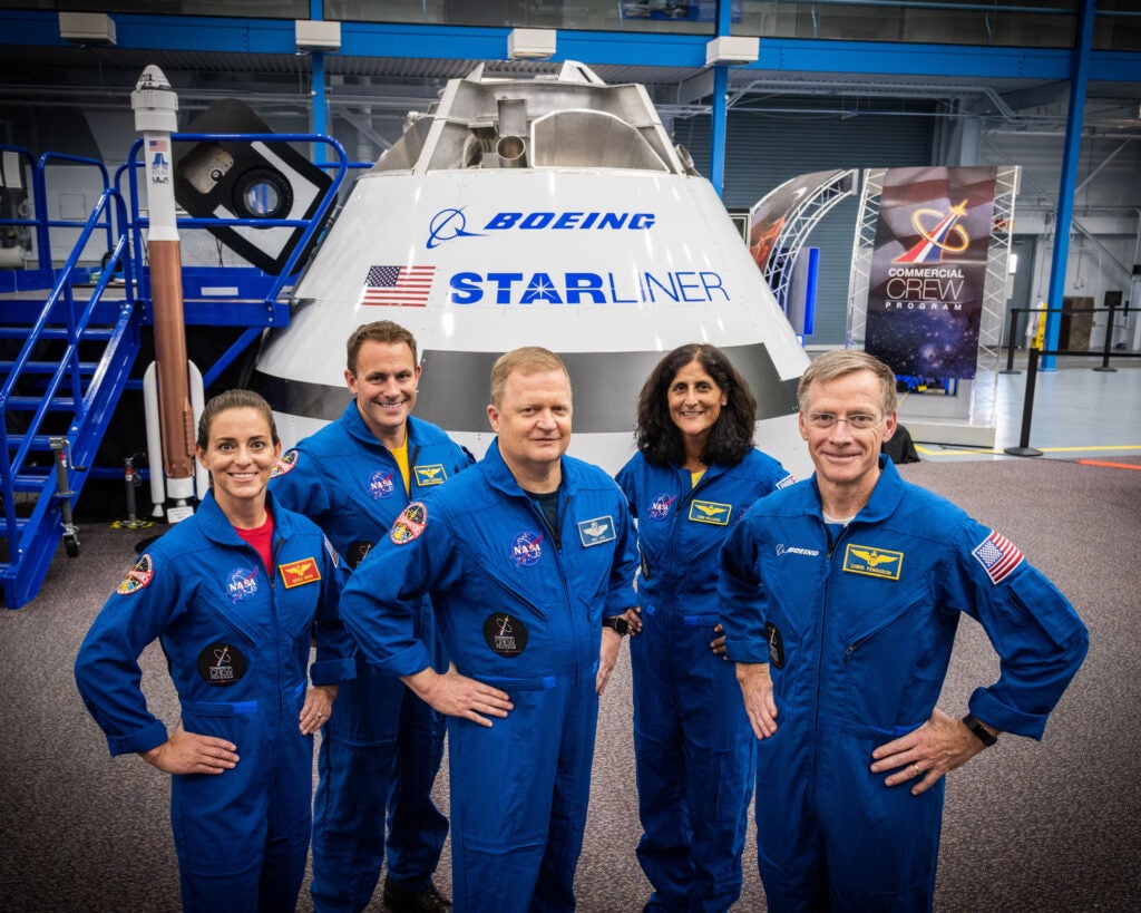 five astronauts in blue jumpsuits standing in front of Starliner capsule