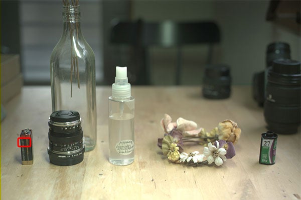 Some bottles, a camera lens, and other objects on a light-colored wooden table.