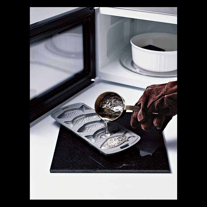 A person wearing thick leather gloves and pouring molten tin solder into a fish shaped mold outside of a microwave.