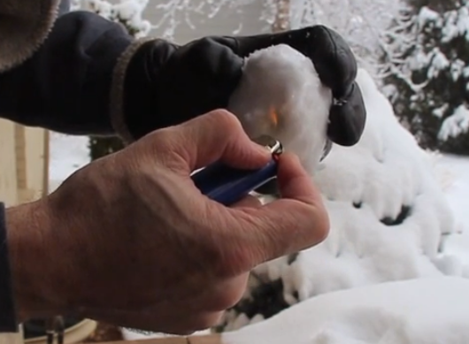 A gloved hand putting a lighter to a snowball