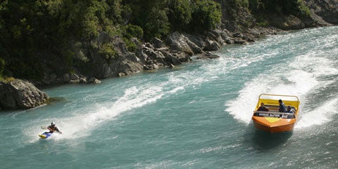 Shaun Baker, left, riding his jet ski-powered kayak on the blue-green waters of New Zealand's Waimakariri River, with a yellow speedboard to his right.