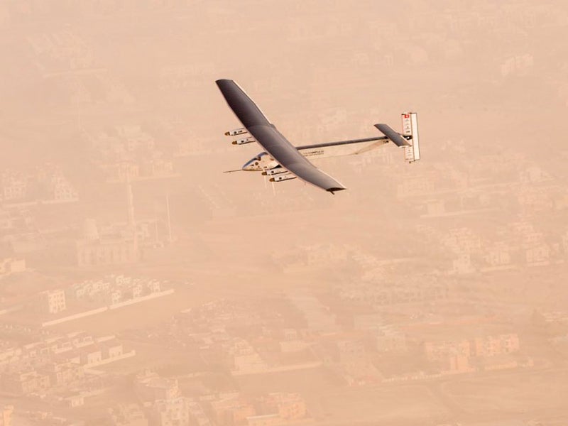 A small glider-style plane with solar panels on the tops of its wings, flying over a dusty urban desert landscape