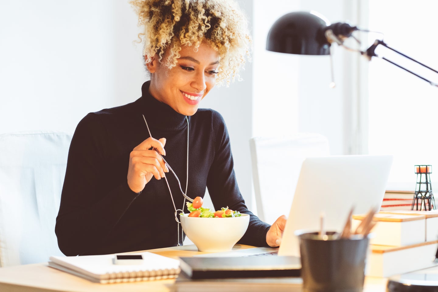 Beautiful afro american young woman sitting at the desk in an office and eating lunch, using laptop at the same time.