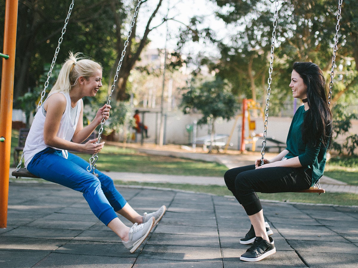 Две женщины сидят лицом друг к другу на качелях на детской площадке и разговаривают друг с другом.  Одна блондинка в джинсах и белой рубашке, другая брюнетка в черных брюках и зеленой рубашке.