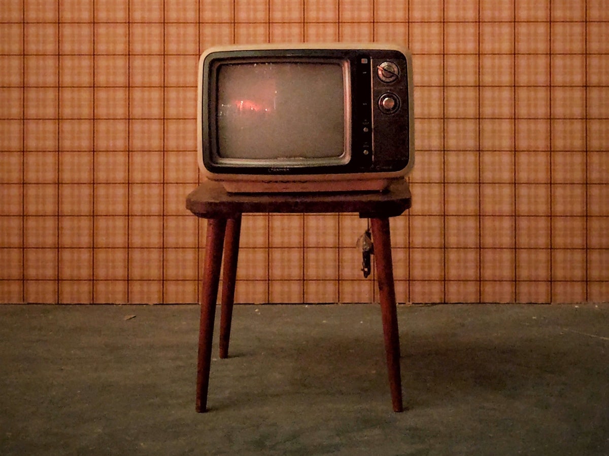 An old TV on a wooden table in front of a wall covered in orange square-patterned wallpaper.