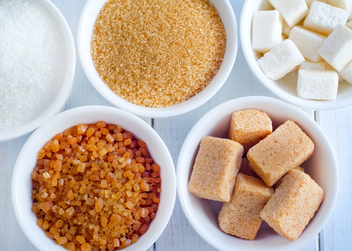 White bowls of white sugar, brown sugar, cane sugar, and unrefined sugar