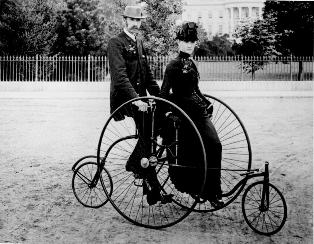 a couple in victorian clothing on an old-fashioned bicycle