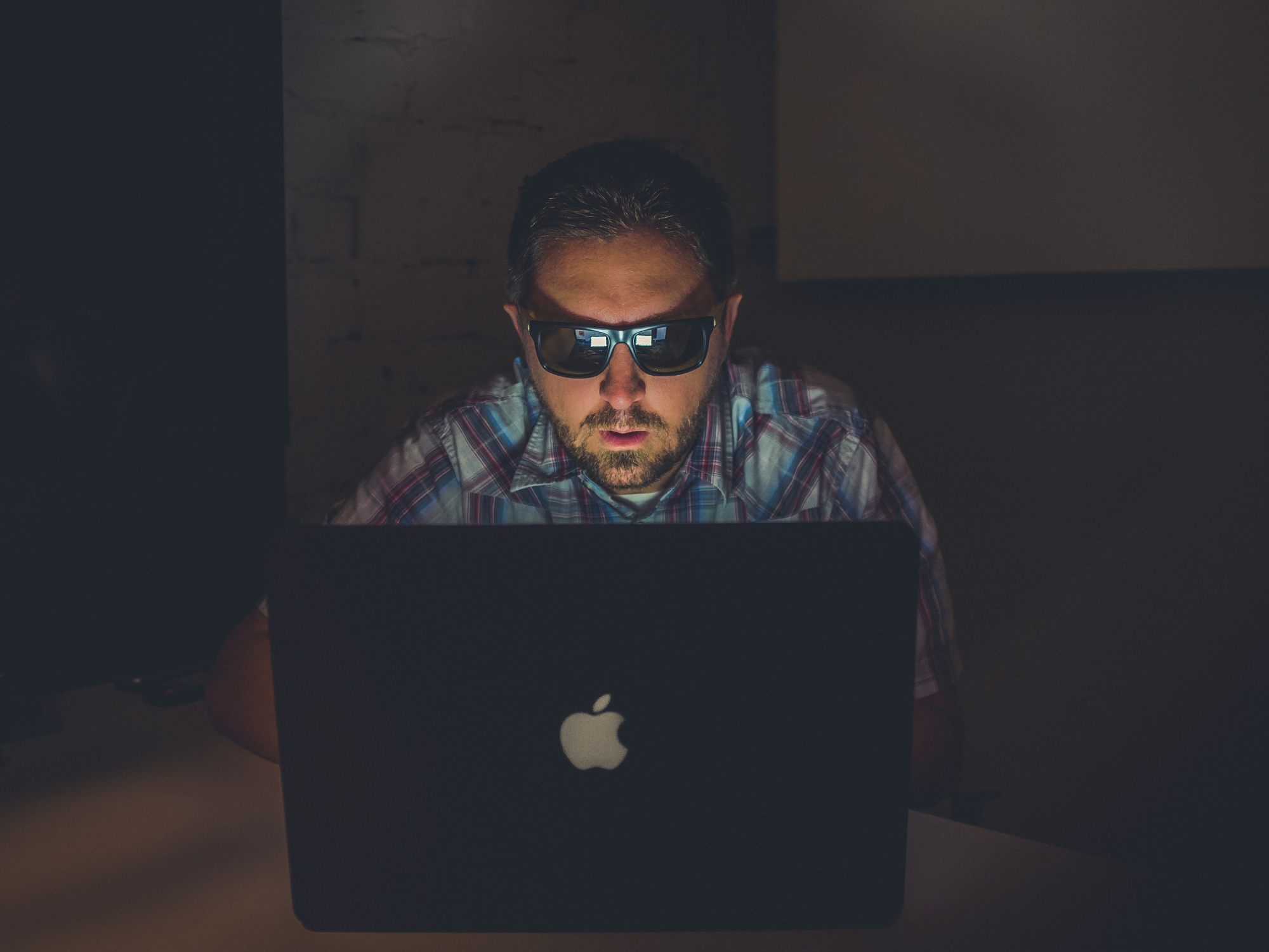 A man wearing sunglasses and a blue plaid shirt sitting in a dark room using an Apple Macbook laptop.