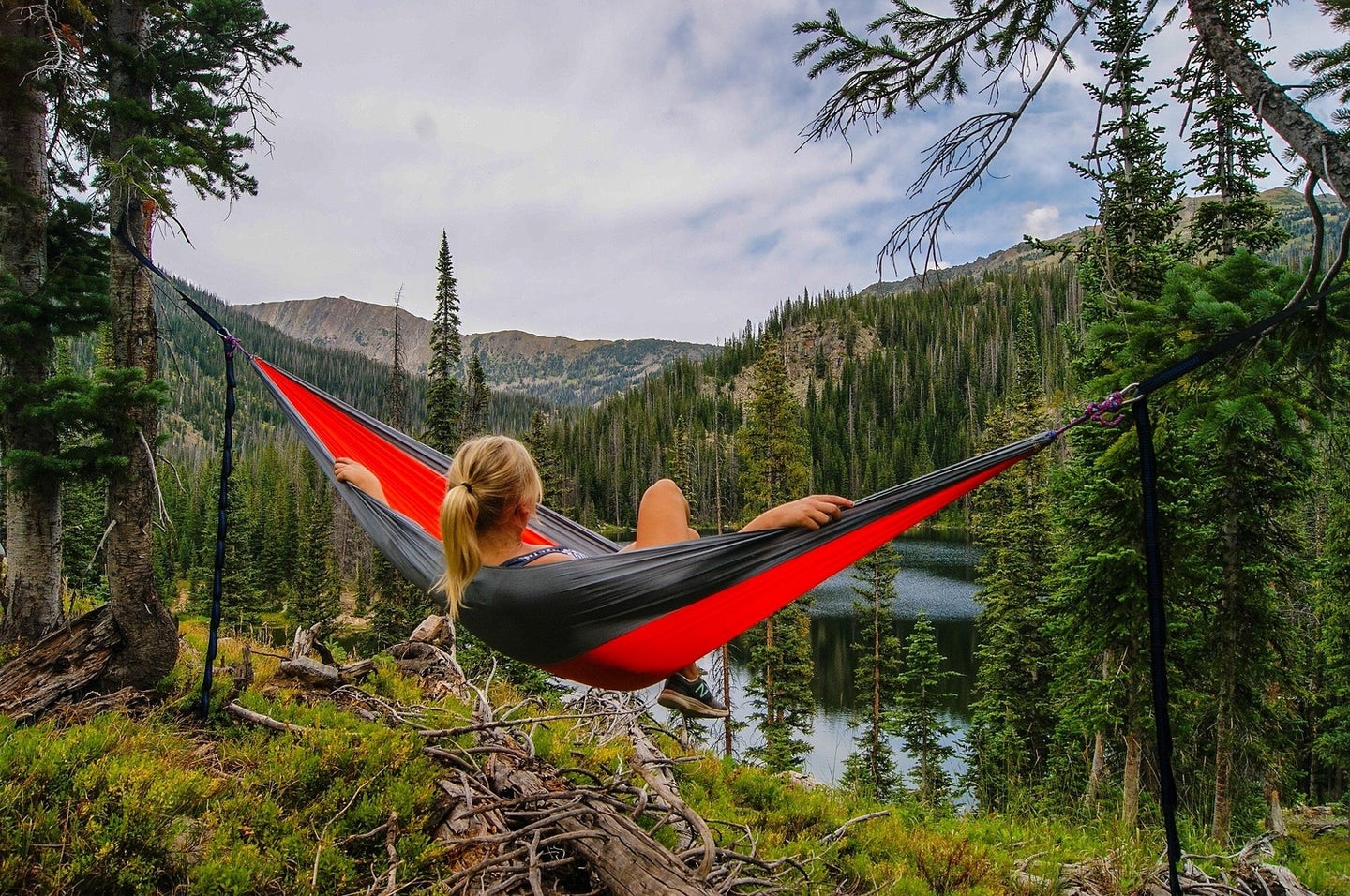A blonde person sitting in a red camping hammock atop a forested ridge, looking out over a lake in some mountains.