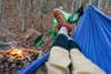 The point of view of a person in a blue hammock near a campfire in the woods, showing their feet and legs from the knees down.