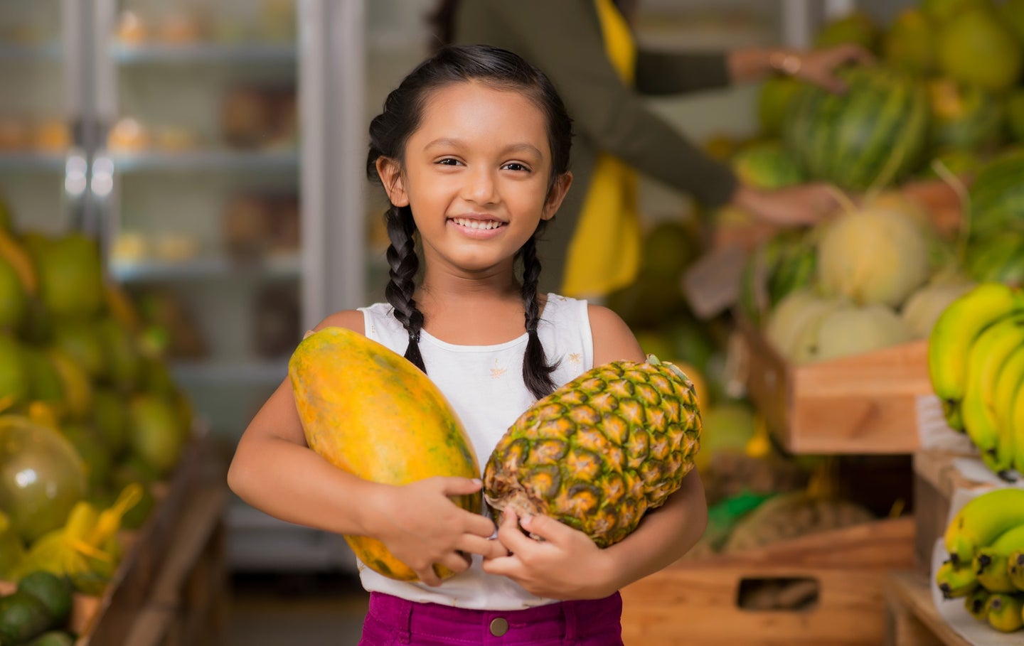 Brown-skinned child with black braids at the grocery store holding ripe fruit like pineapple and papaya in arms
