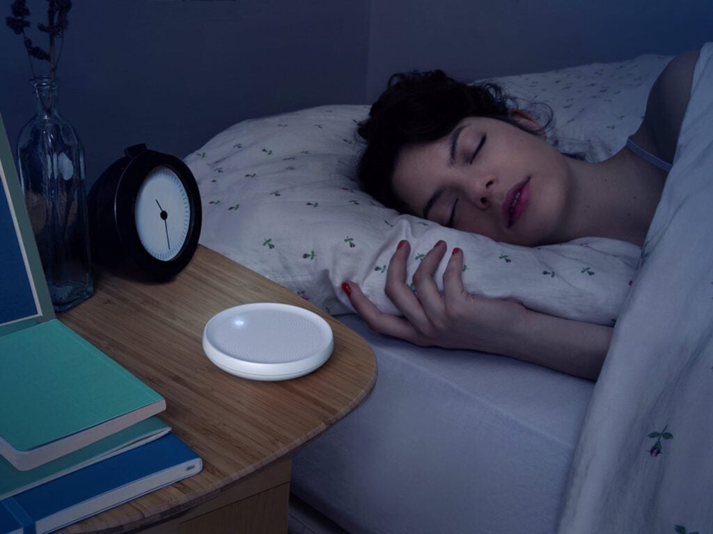 A woman sleeping in a bed with a Dodow Sleep Aid device next to her on the bedside table.