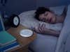 A woman sleeping in a bed with a Dodow Sleep Aid device next to her on the bedside table.