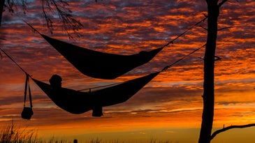 A person in a hammock between two trees, with another hammock above him, on a shoreline at sunset.