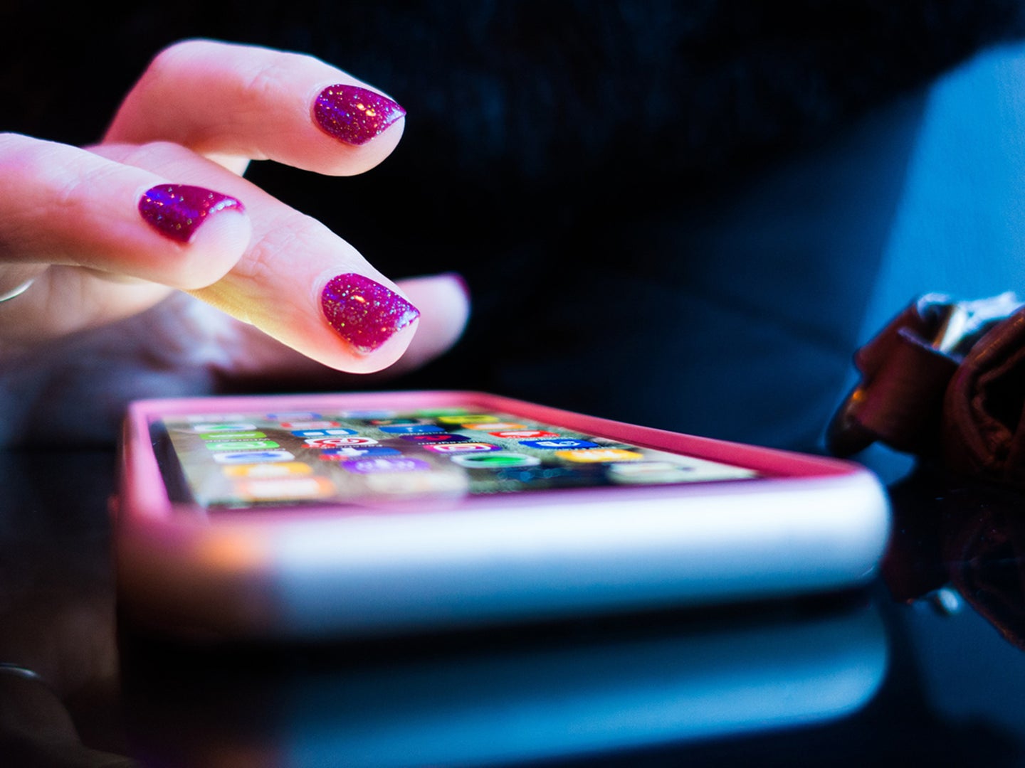 A hand with magenta painted nails reaches out from the darkness to tap on a phone lying on a shiny black table.