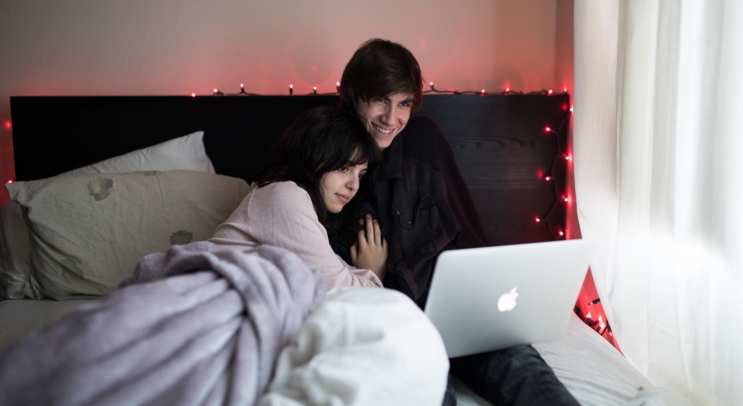 Two people cuddling in bed while watching Netflix or something else on a silver Macbook laptop.
