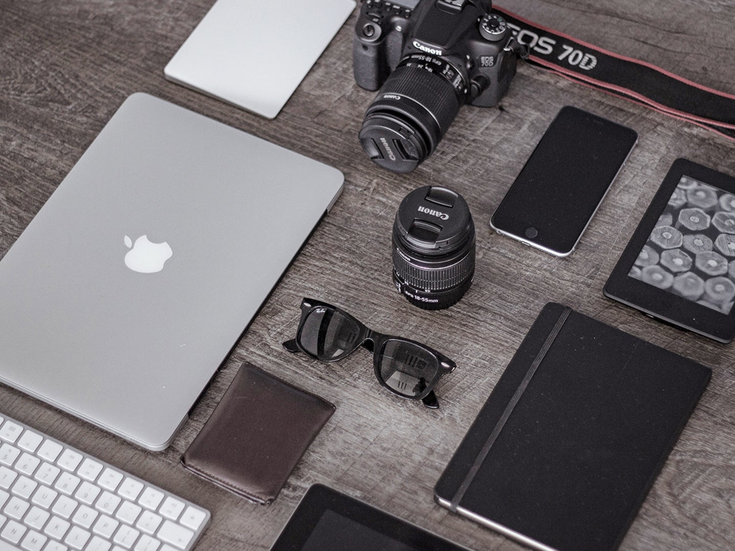 A MacBook laptop, an Apple keyboard, a digital camera, an iPhone, a camera lens, sunglasses, an iPad, and some notebooks on a gray wooden table.