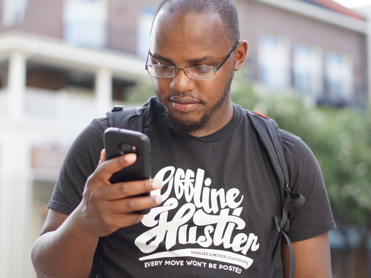 A Black man wearing a t-shirt, a backpack, and glasses, looking annoyed at a spam call on his cell phone.