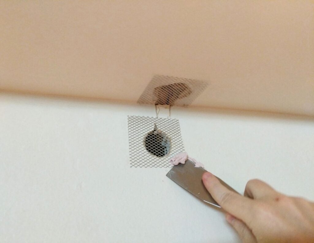 a person applying putty with a putty knife to a wire mesh patch over a hole in drywall