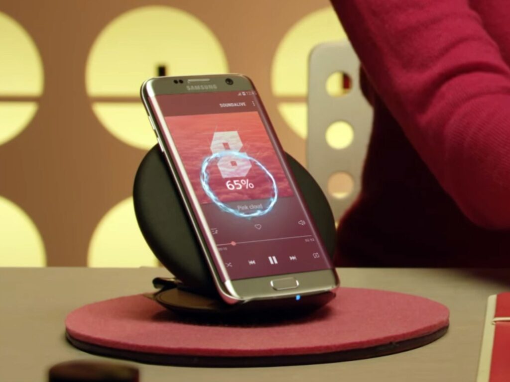 A black Samsung phone recharging on a black wireless charging pad that's sitting on a red platform on a tan table.