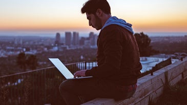 A man working on a laptop while sitting on a rock overlooking a city at sunset.