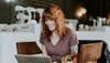 A redheaded woman in a coffee shop smiling while she uses a laptop.